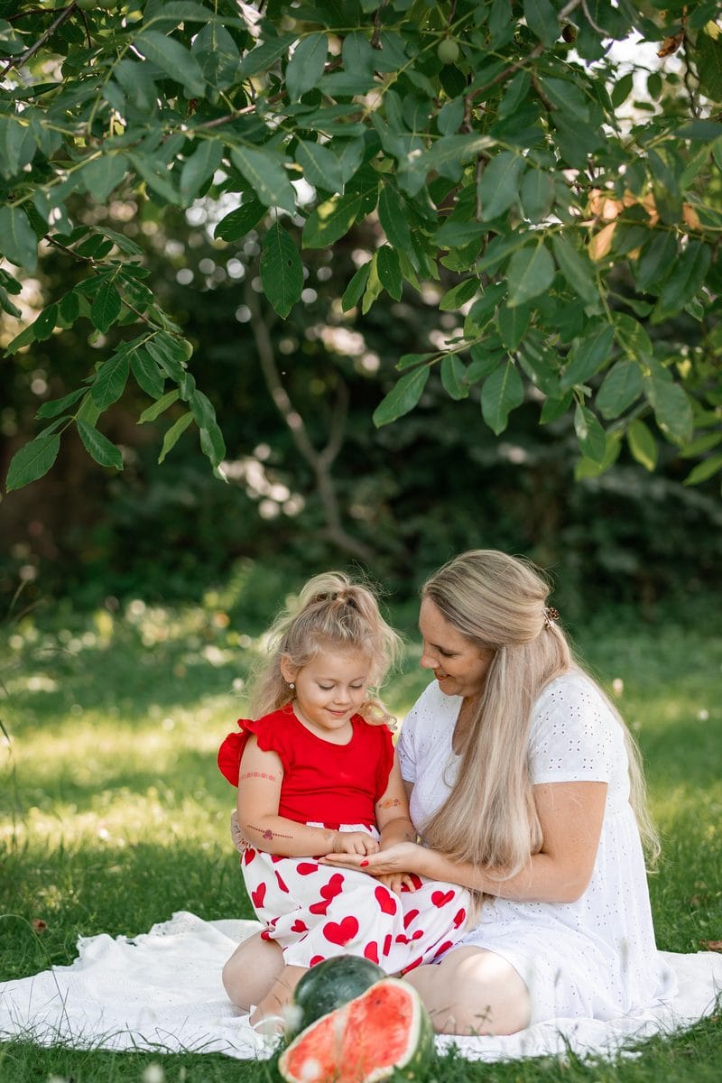 Maminka s dcerkou sedí na bílé dece pod stromem, před nimi rozkrojený meloun