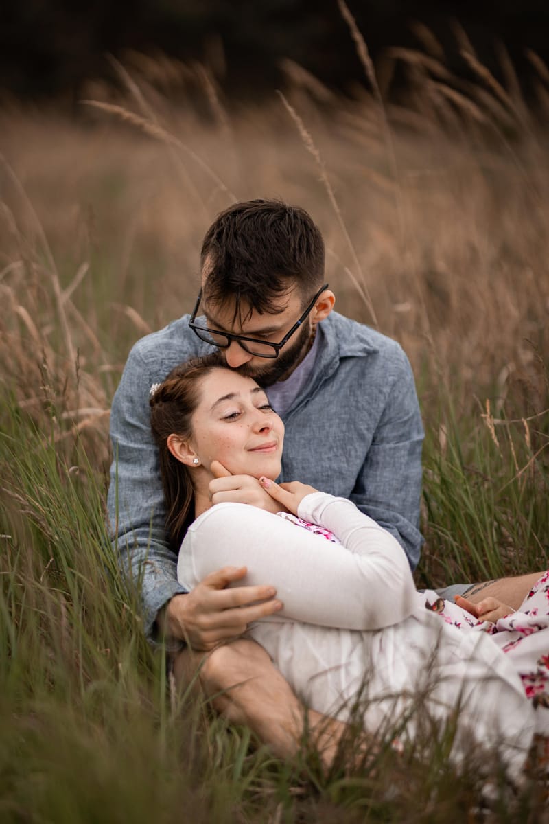Žena se zavřenýma očima se opírá o partnera, který ji líbá do vlasů, sedí ve vysoké trávě