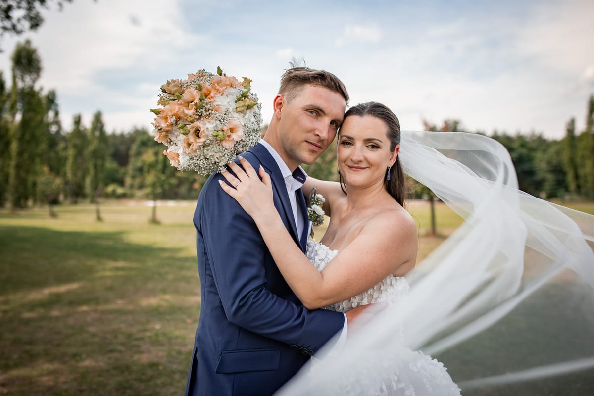 Novomanželé v objetí se usmívají do objektivu, nevěsta drží kytici a vítr jí zvedá závoj