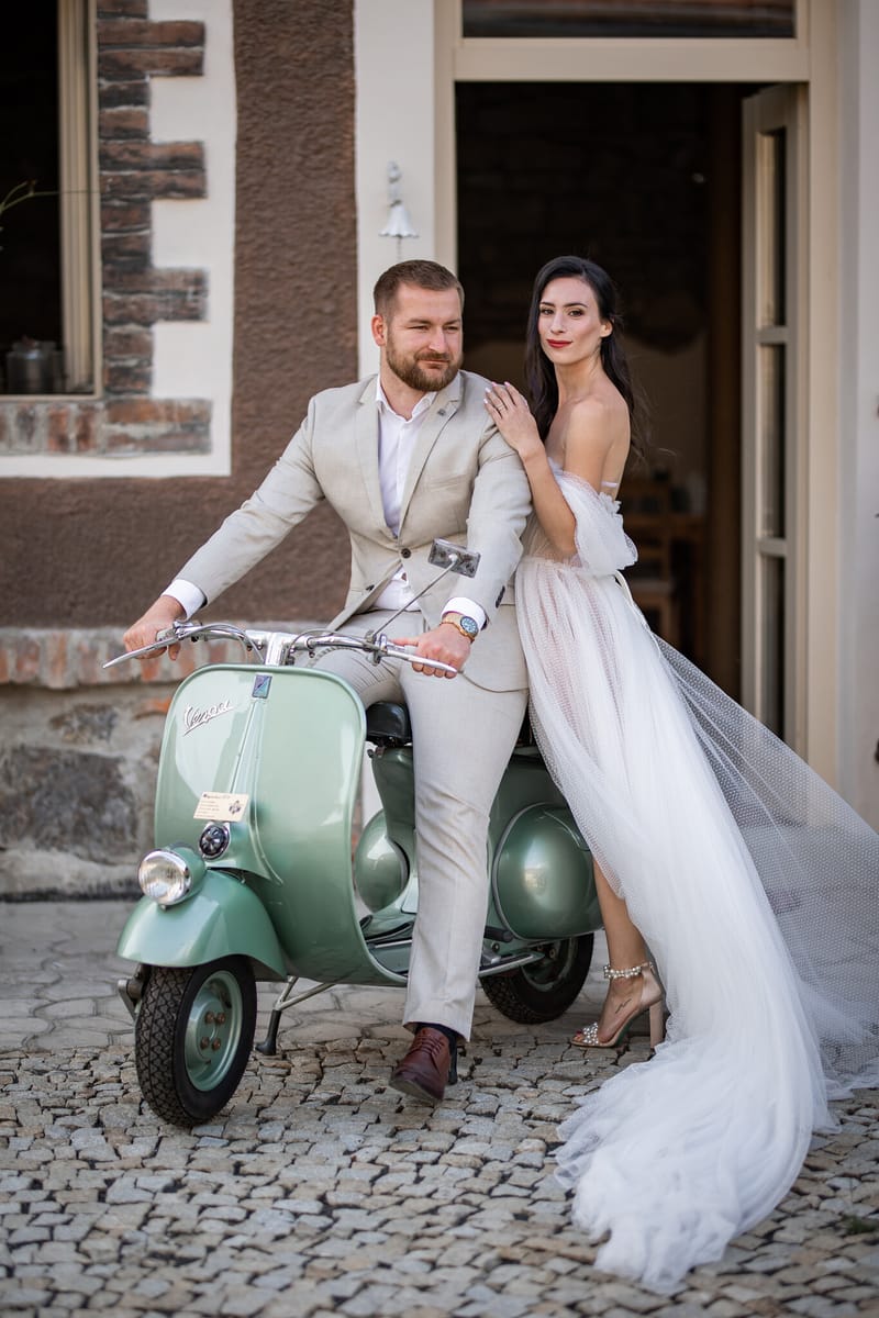 Ženich ve světlém obleku sedí na zelené vespě a nevěsta v tylových šatech se k němu tiskne