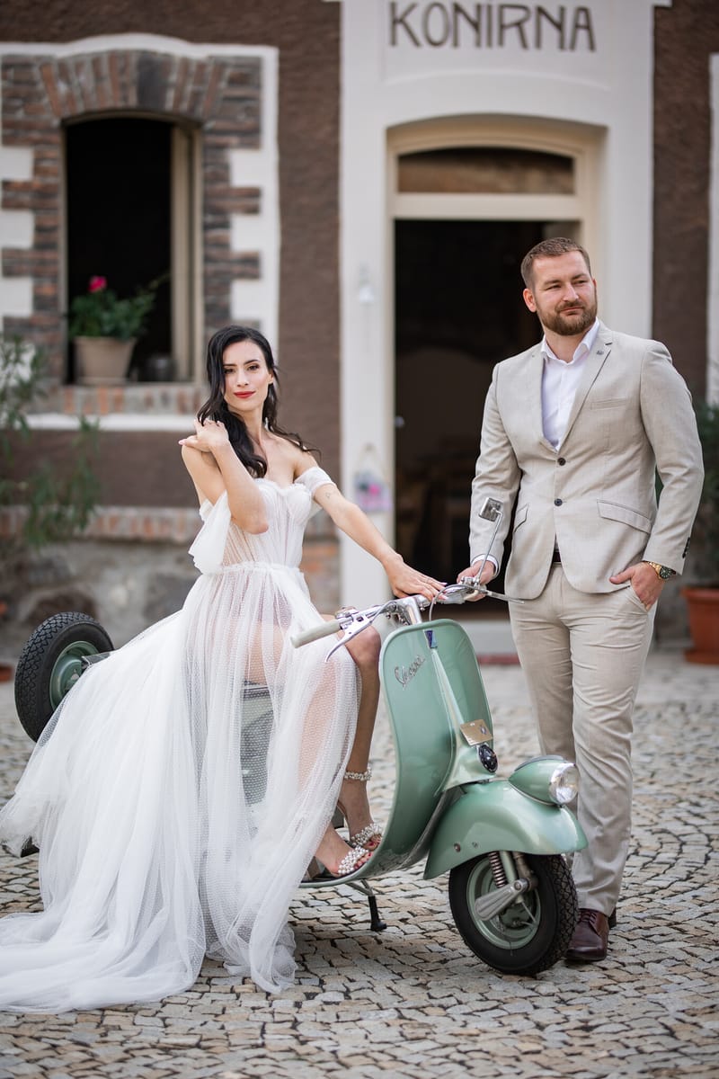 Nevěsta v tylových šatech sedí na zelené vespě a ženich ve světlém obleku stojí vedle ní