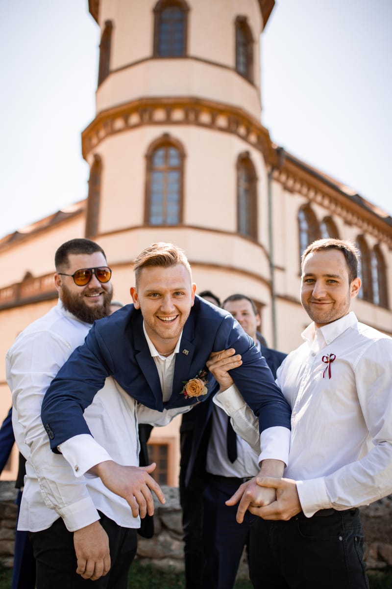 Rozesmátý ženich v náručí svědků před zámeckou věží za slunečného dne