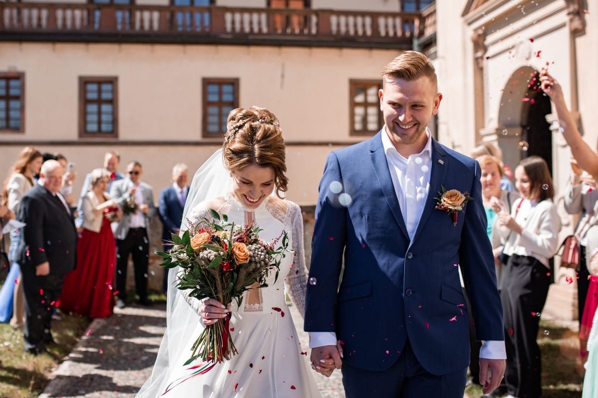 Novomanželé s úsměvem odcházejí od obřadu, hosté na ně hází okvětní lístky