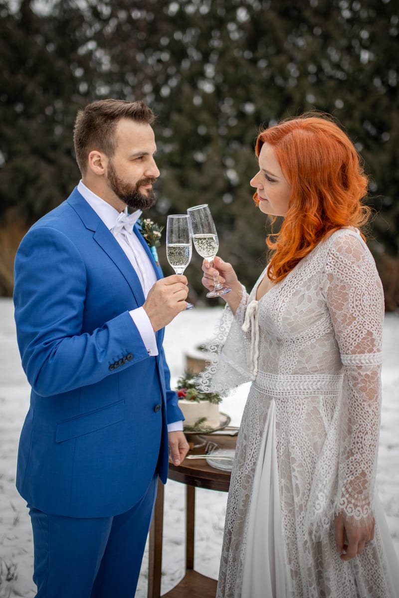 Ženich v modrém obleku a nevěsta v krajkových šatech si připíjejí šampaňským ve sněhu