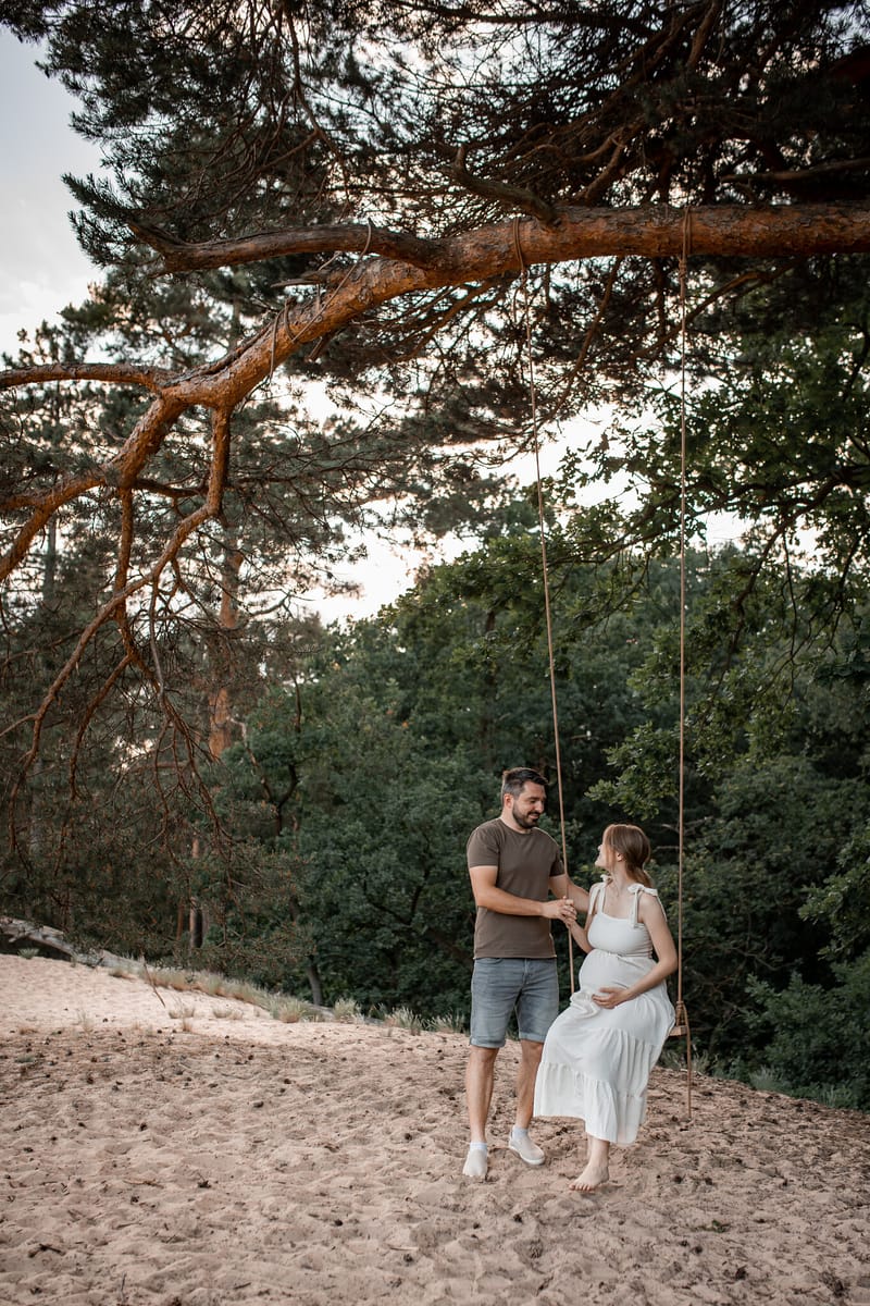 Těhotná maminka sedí na houpačce pod velkou borovicí, partner stojí vedle ní na písku