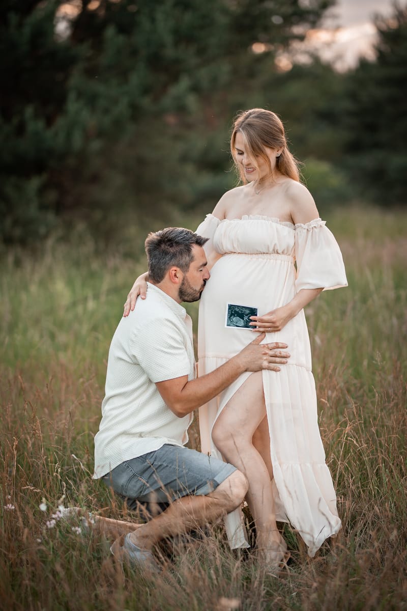 Partner klečí ve vysoké trávě a líbá bříško, maminka drží fotku z ultrazvuku