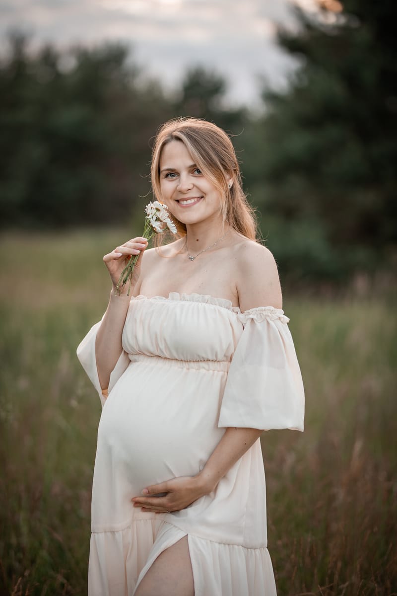 Těhotná maminka v krémových šatech drží luční kvítek a hladí si bříško na louce