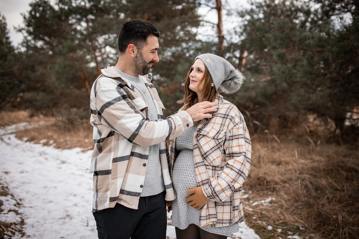 Partner upravuje těhotné mamince límec kabátu, usmívají se na sebe na zasněžené cestě