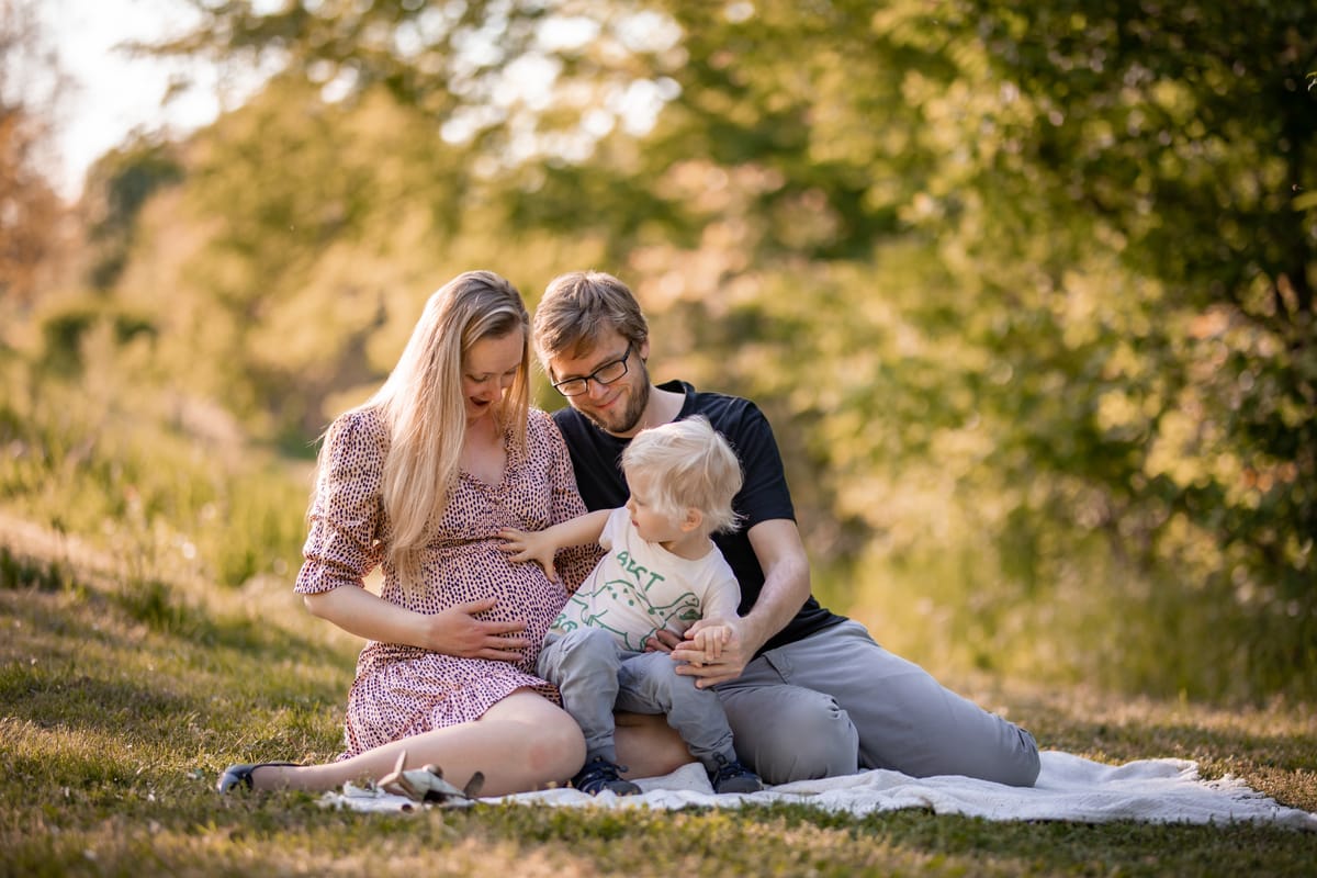Těhotná maminka, tatínek a malý chlapeček sedí na dece v trávě, chlapeček sahá na bříško, zlaté světlo