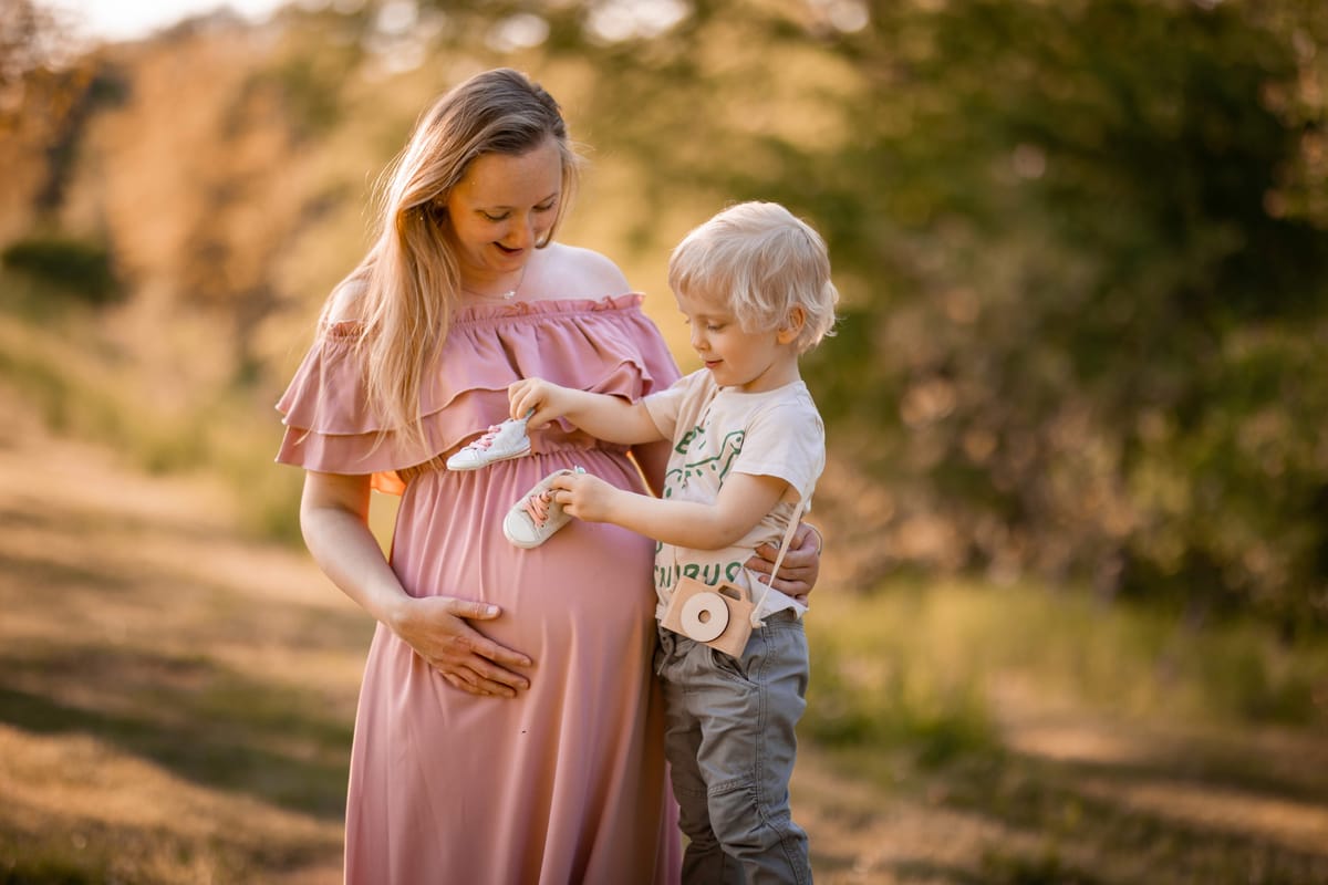 Chlapeček přikládá dětské botičky k bříšku usměvavé těhotné maminky v růžových šatech na louce